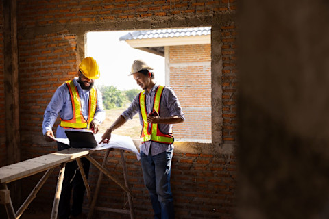 Two workers wearing collared shirts, work vests and hard hats look over architectural plans on a construction site in a room with an old brick wall.