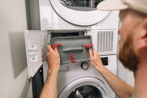 A person services a modern washing machine and dryer unit. The appliance doors and control panels are visible.