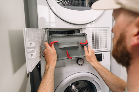 A person services a modern washing machine and dryer unit. The appliance doors and control panels are visible.