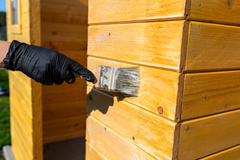 Closeup on a worker’s gloved hand applying finish to a wooden garden shed.